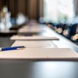 Close up of dark conference table with water glasses, pens, paper sheets and blurry window background