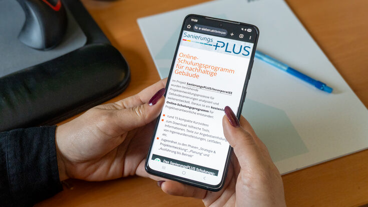 Frau hält Mobiltelefon in der Hand, wo am Bildschirm das Online-Schulungsprogramm für nachhaltige Gebäude zu sehen ist.