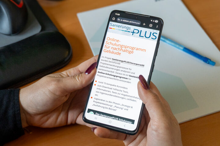 Frau hält Mobiltelefon in der Hand, wo am Bildschirm das Online-Schulungsprogramm für nachhaltige Gebäude zu sehen ist.