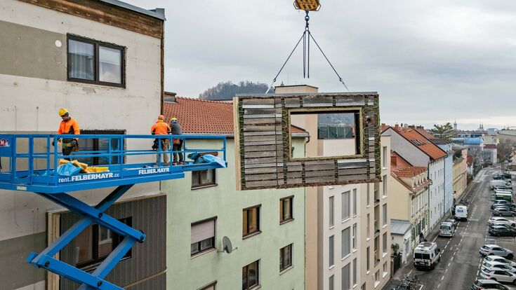 Kran, an dem ein CEPA-Energiefassaden-Element (von TOWERN3000) hängt, das von Monteuren an die Fassade eines Gebäudes angebracht wird.