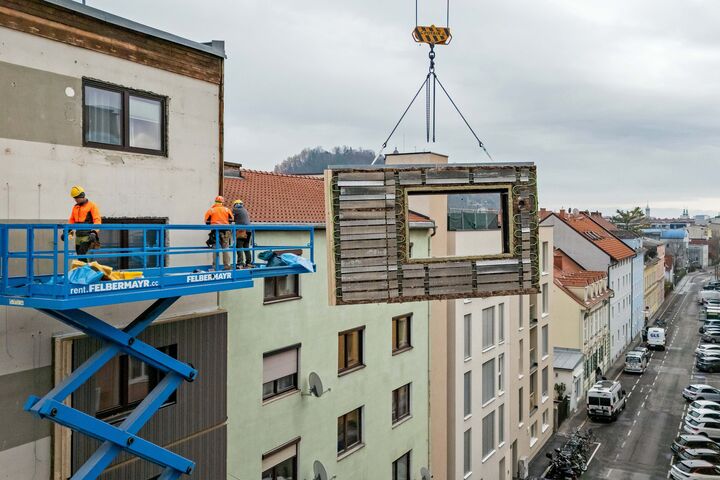 Kran, an dem ein CEPA-Energiefassaden-Element (von TOWERN3000) hängt, das von Monteuren an die Fassade eines Gebäudes angebracht wird.